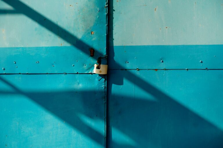 Gros plan d'une porte métallique bleue sécurisée par un cadenas, projetant des ombres au soleil.