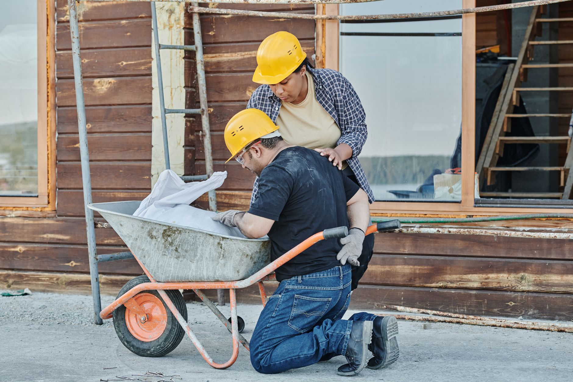 Un ouvrier du bâtiment recevant de l'aide alors qu'il est agenouillé avec une brouette sur un chantier.