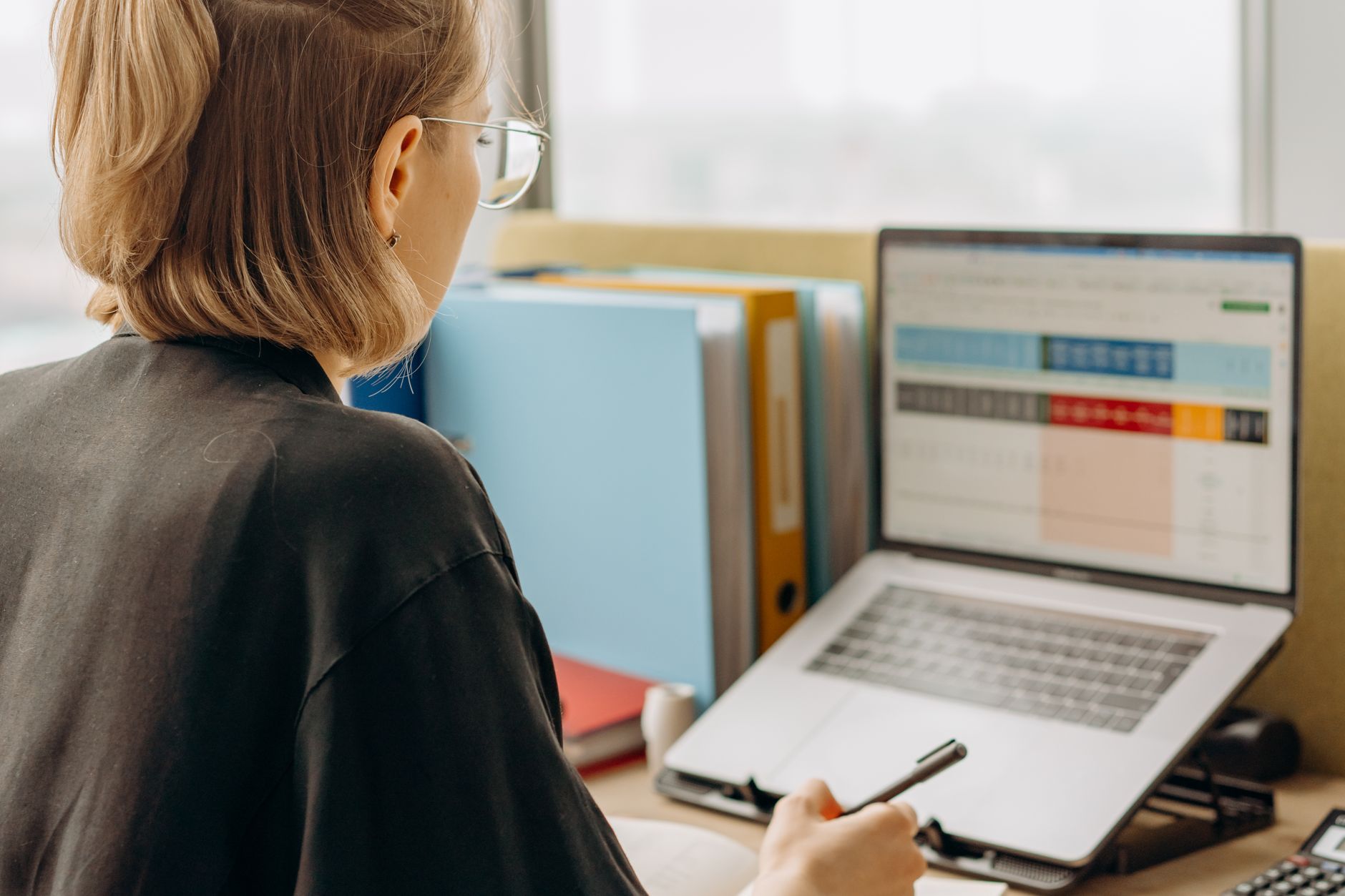Femme de dos devant un ordinateur portable affichant un tableau, un stylo à la main, avec des classeurs et une calculatrice sur le bureau.