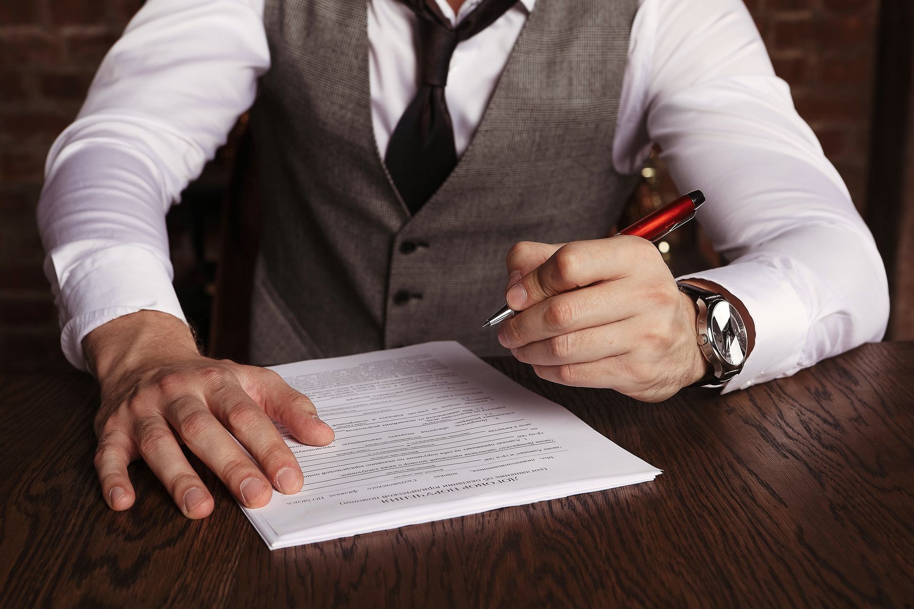 Homme en gilet et cravate, un stylo rouge à la main, prêt à signer un document posé sur une table en bois.