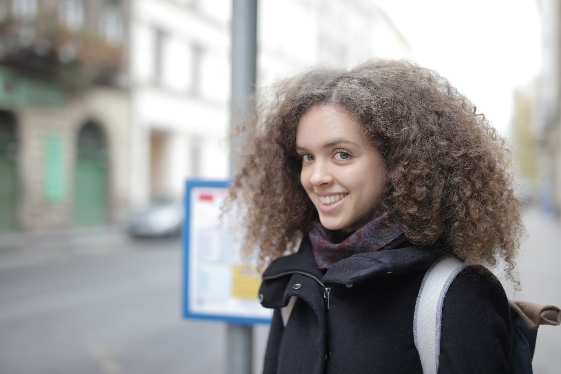 Jeune femme souriante aux cheveux bouclés, manteau sombre et sac à dos, debout dans une rue de ville devant des immeubles flous.