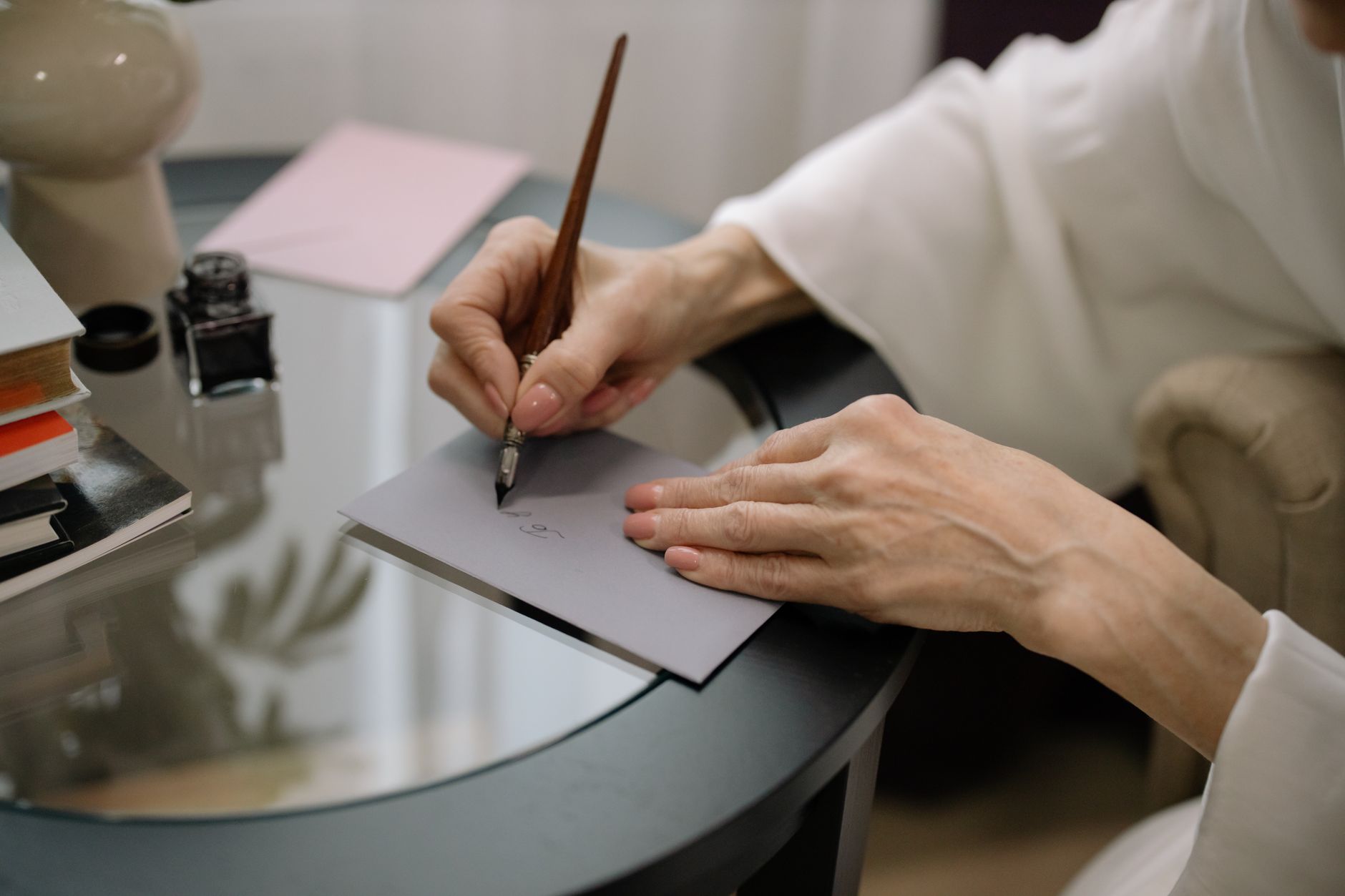Mains d’une personne écrivant à la plume sur une enveloppe grise, sur une table ronde en verre avec un encrier et des livres.