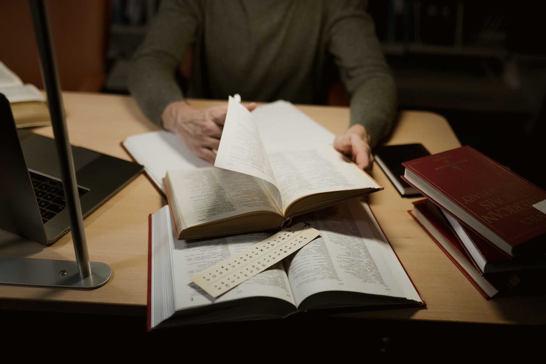 Personne feuilletant un gros livre ouvert sur un bureau, avec des livres rouges empilés, un ordinateur portable et une lampe, dans une ambiance de bibliothèque.