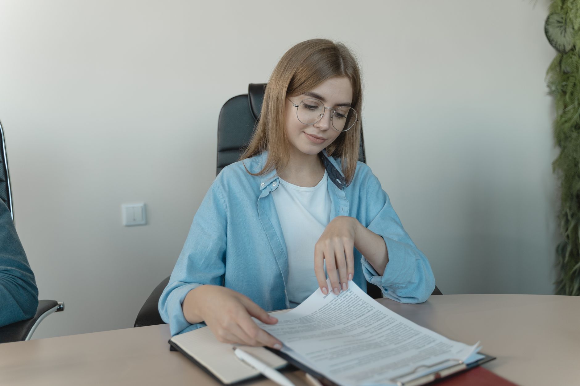 Une femme à lunettes en chemise bleue, assise à une table de bureau, feuillette des documents posés sur un porte-bloc.