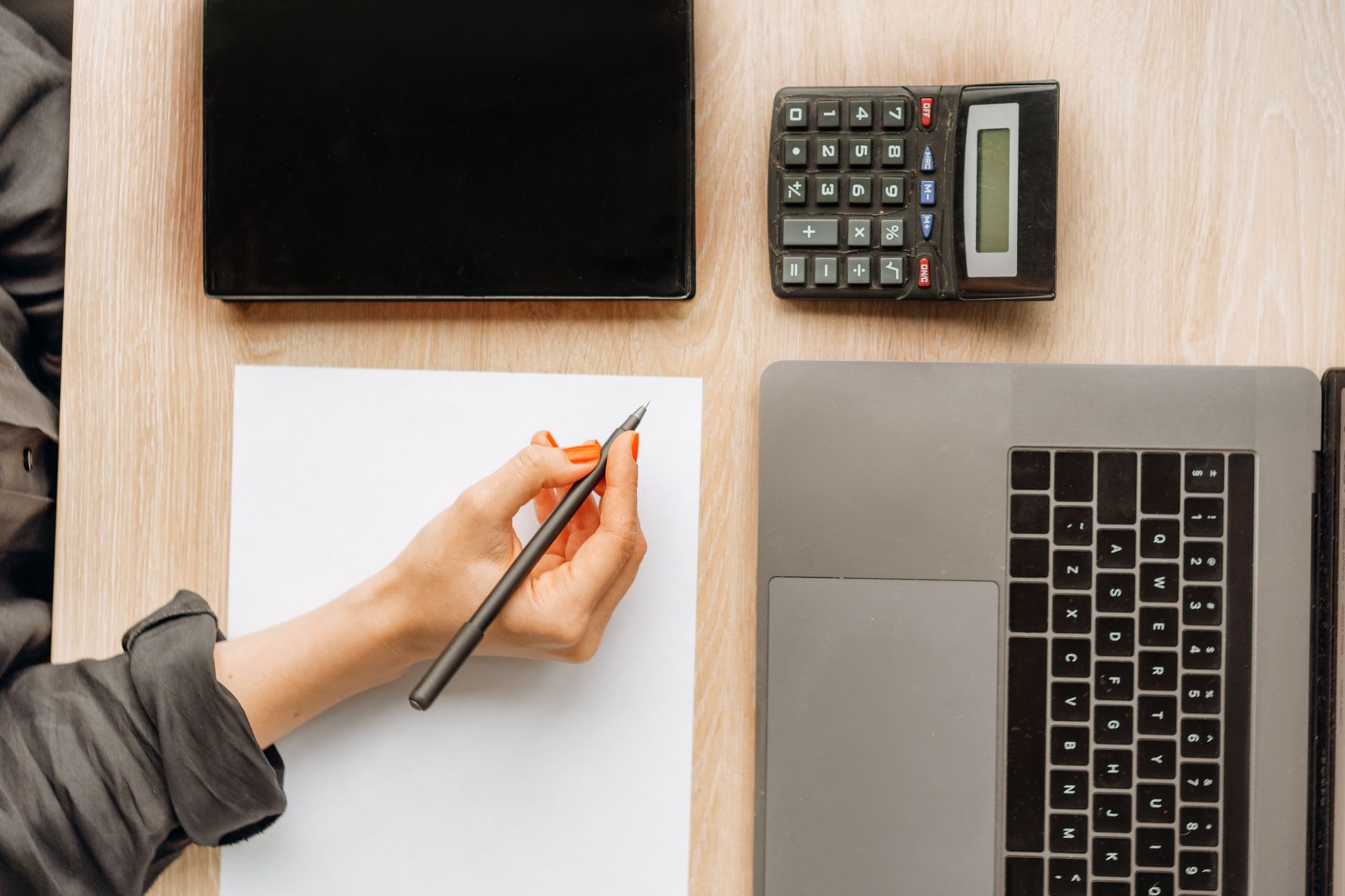 Vue de dessus d’un bureau : une main tenant un stylo sur une feuille vierge, avec une calculatrice, un ordinateur portable et un carnet noir.